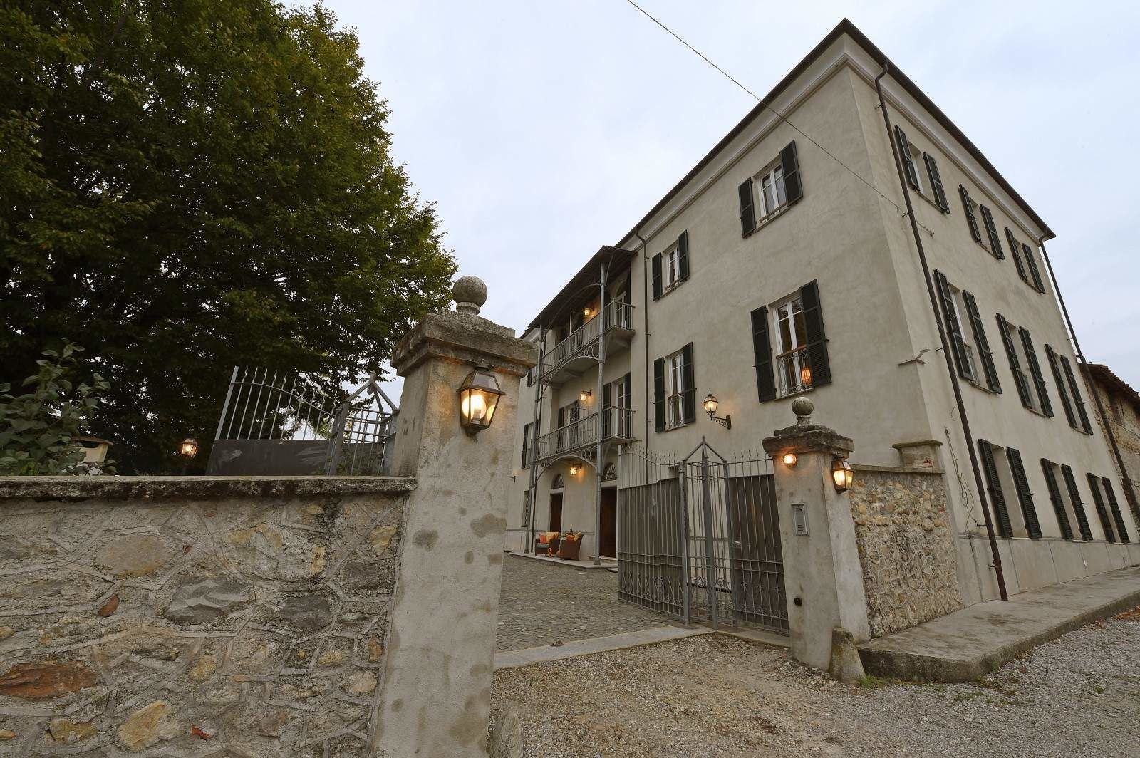 A two-story building with a stone wall entrance, a metal gate, and a large tree to the left.