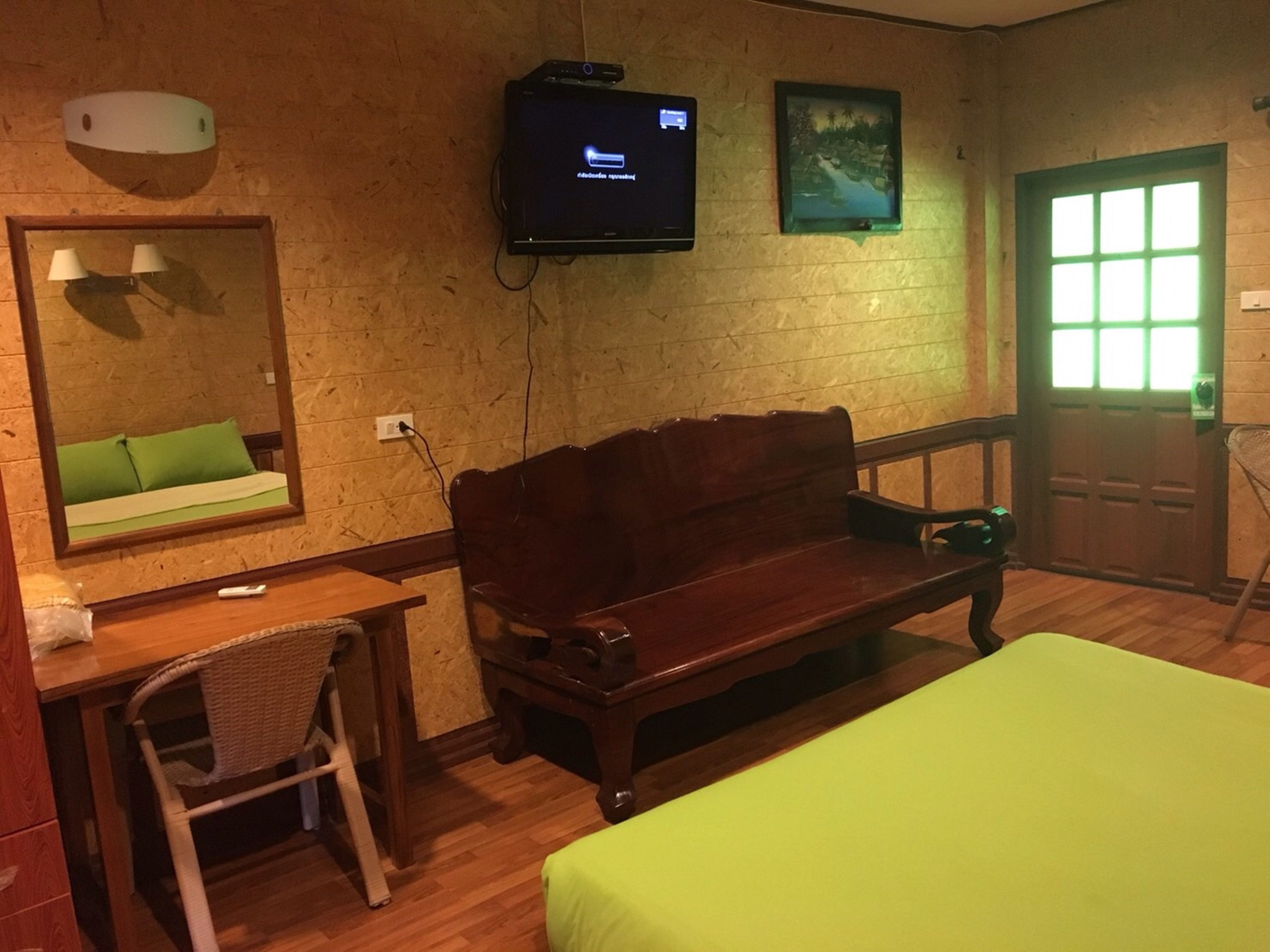 Una habitación con un banco de madera, un escritorio con una silla, una cama, un televisor montado en la pared y una ventana con una puerta iluminada en verde.