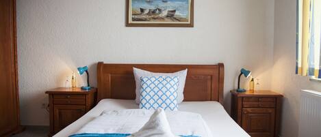A hotel room with a bed, a painting of boats on the wall, two bedside tables with lamps, and a window with curtains.