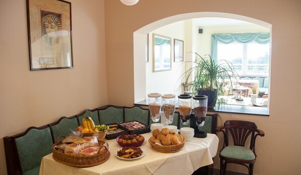 A dining area with a table set for breakfast, including bread, pastries, and fruit. There are also coffee and tea dispensers on the table. - Hotel Atlantis (Ramstein-Miesenbach)