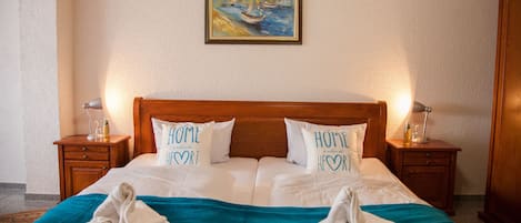 Habitación de hotel con cabecera de madera, dos mesitas de noche con lámparas, un cuadro de barcos en la pared y almohadas con la inscripción 'HOME is where the HEART is'.