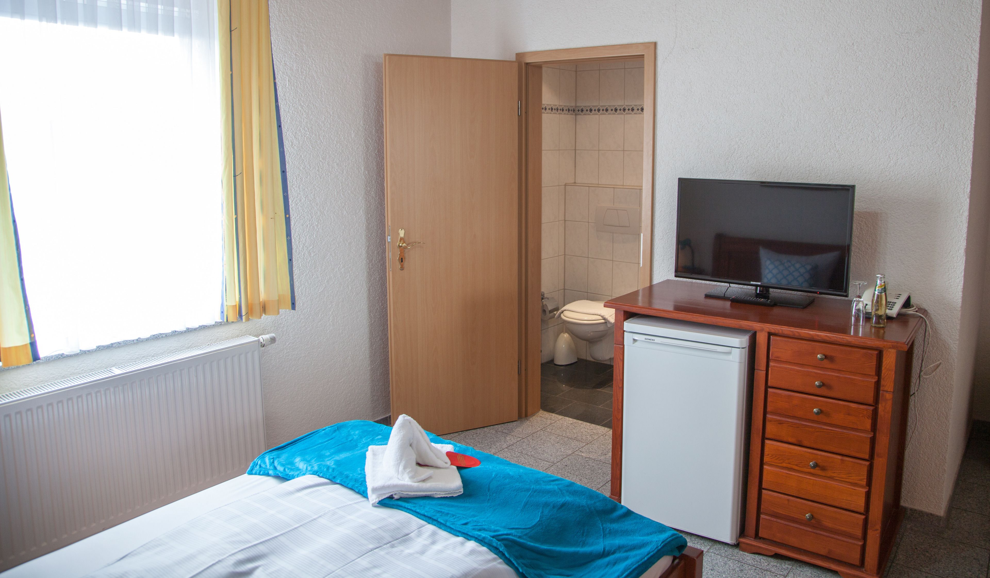 A hotel room with a bed, a small desk with a television, a refrigerator, and a bathroom visible through an open door.