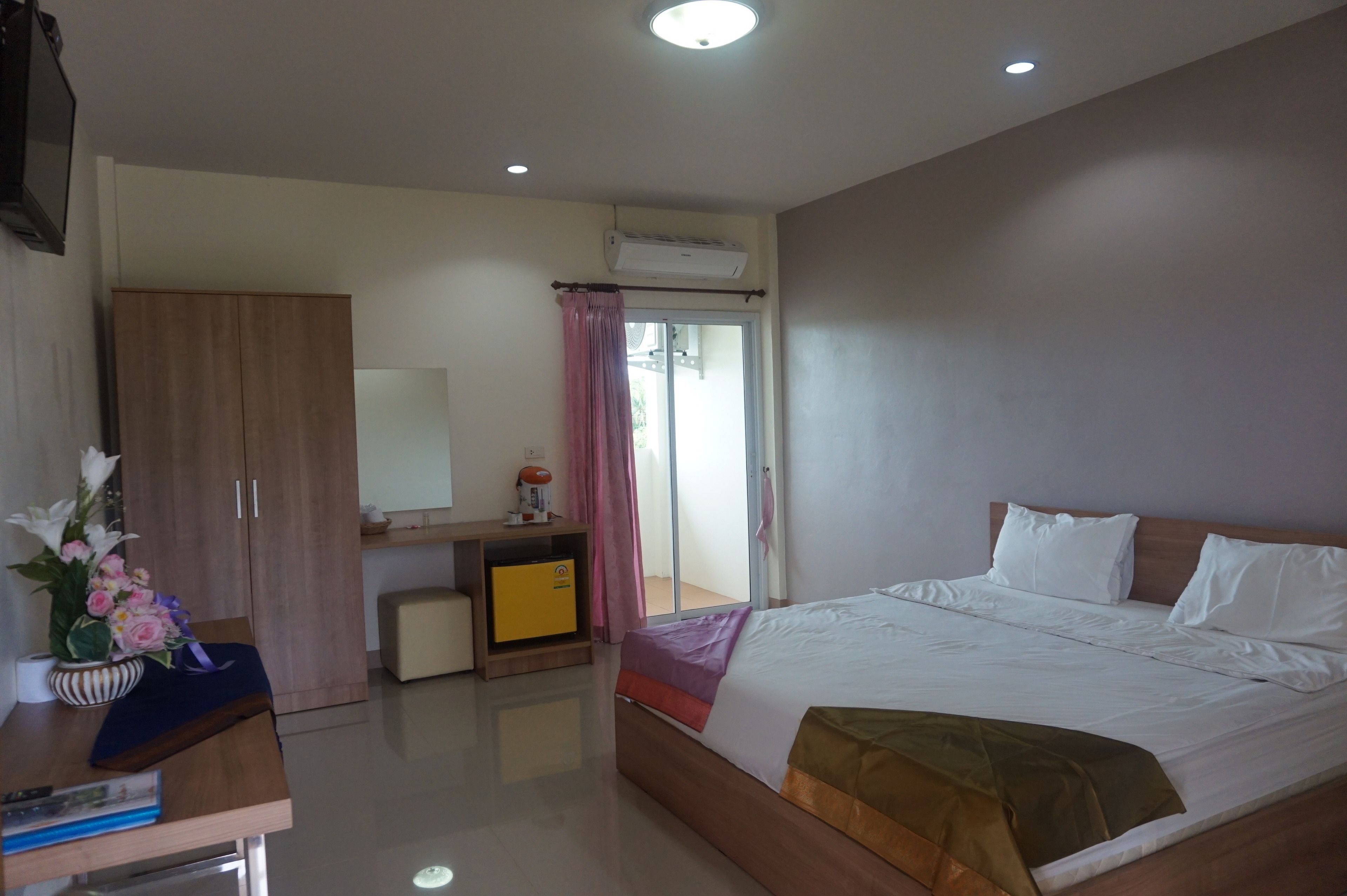 A hotel room with a bed, a desk with a chair, a wardrobe, a mirror, an air conditioning unit, and a window with curtains.