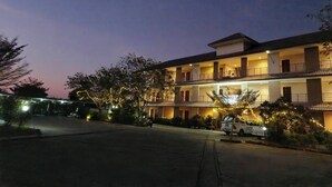 A multi-story hotel with a balcony on the second floor, a parking area, and trees with lights. - Bumnsaya Place (Nang Rong)