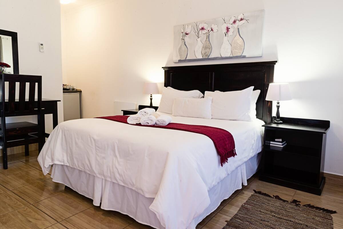 A neatly made bed with white bedding and a red throw, a wooden chair, a bedside table with a lamp, and a wall-mounted artwork of vases with flowers.