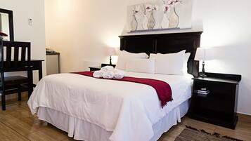 A neatly made bed with white bedding and a red throw, a wooden chair, a bedside table with a lamp, and a wall-mounted artwork of vases with flowers.