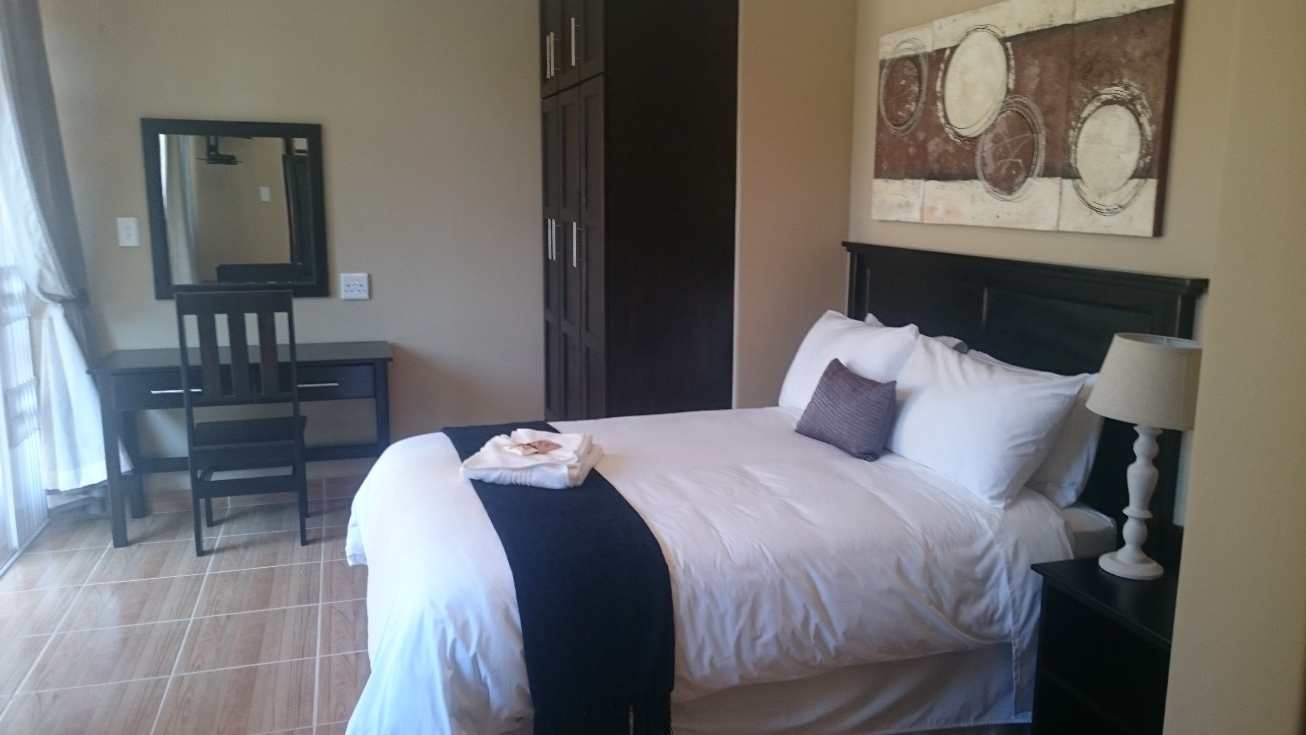 A hotel room with a bed, a desk, a chair, a lamp, and a painting on the wall.