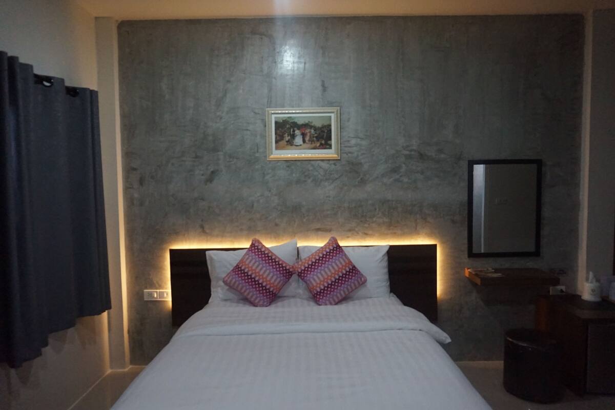 A bedroom with a bed, two decorative pillows, a framed picture on the wall, a nightstand, and a mirror.