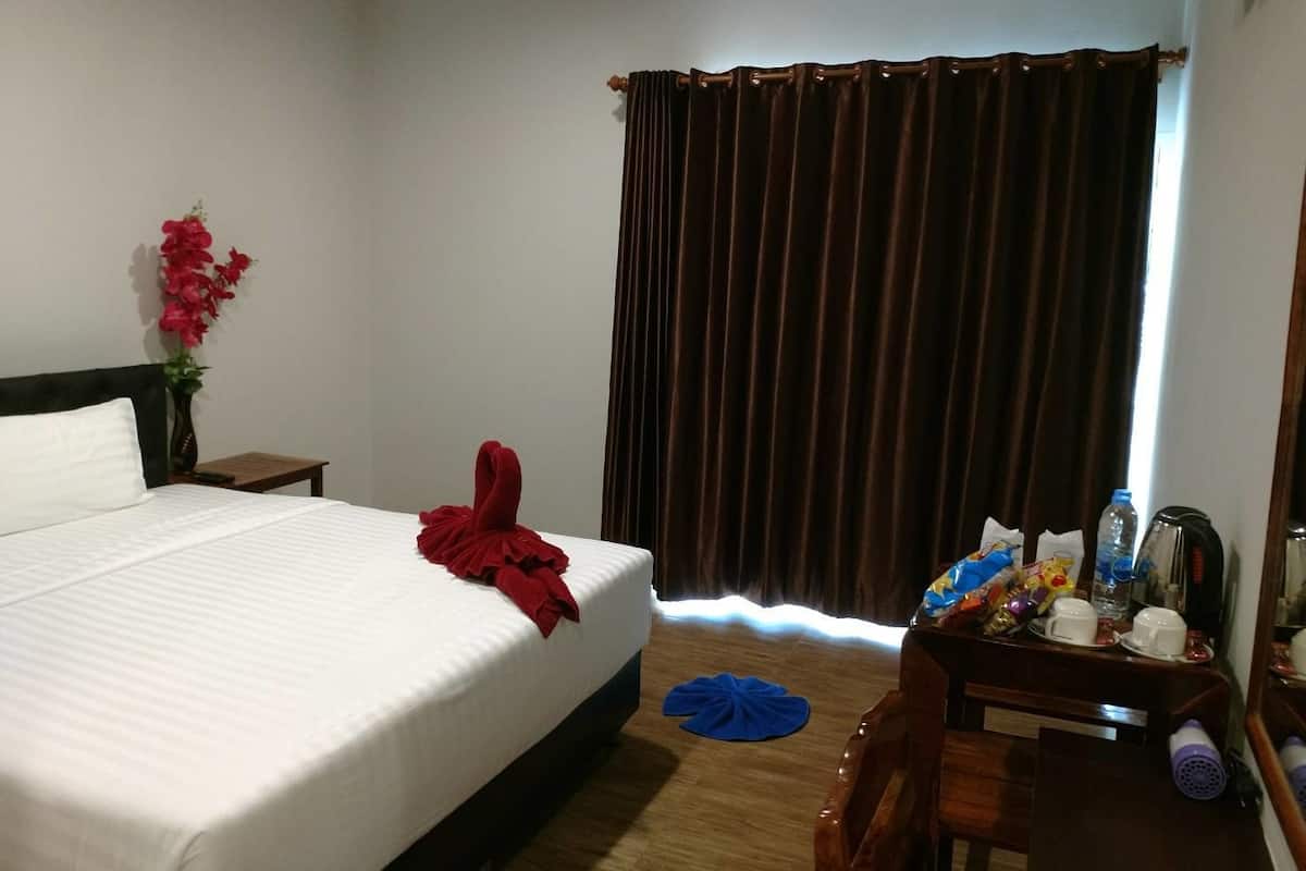 A hotel room with a bed, a nightstand, a desk with a chair, a mirror, a window with curtains, and a vase with flowers.
