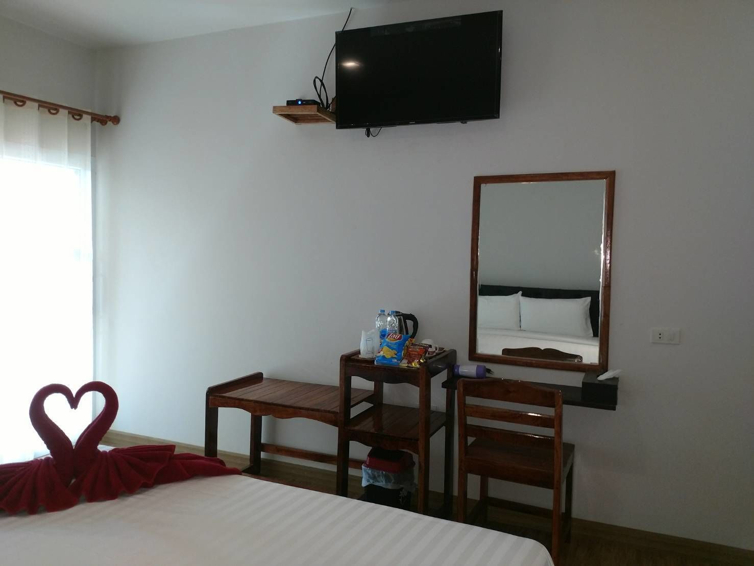A hotel room with a bed, a desk with a chair, a television mounted on the wall, and a mirror.