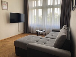 Una sala de estar moderna con un sofá seccional gris, un televisor de pantalla plana montado en la pared, una pequeña mesa redonda y cortinas translúcidas que dejan entrar la luz natural.