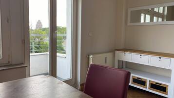 A room with a wooden table, a red chair, a white cabinet with drawers, and a window with a view of greenery and a building outside.