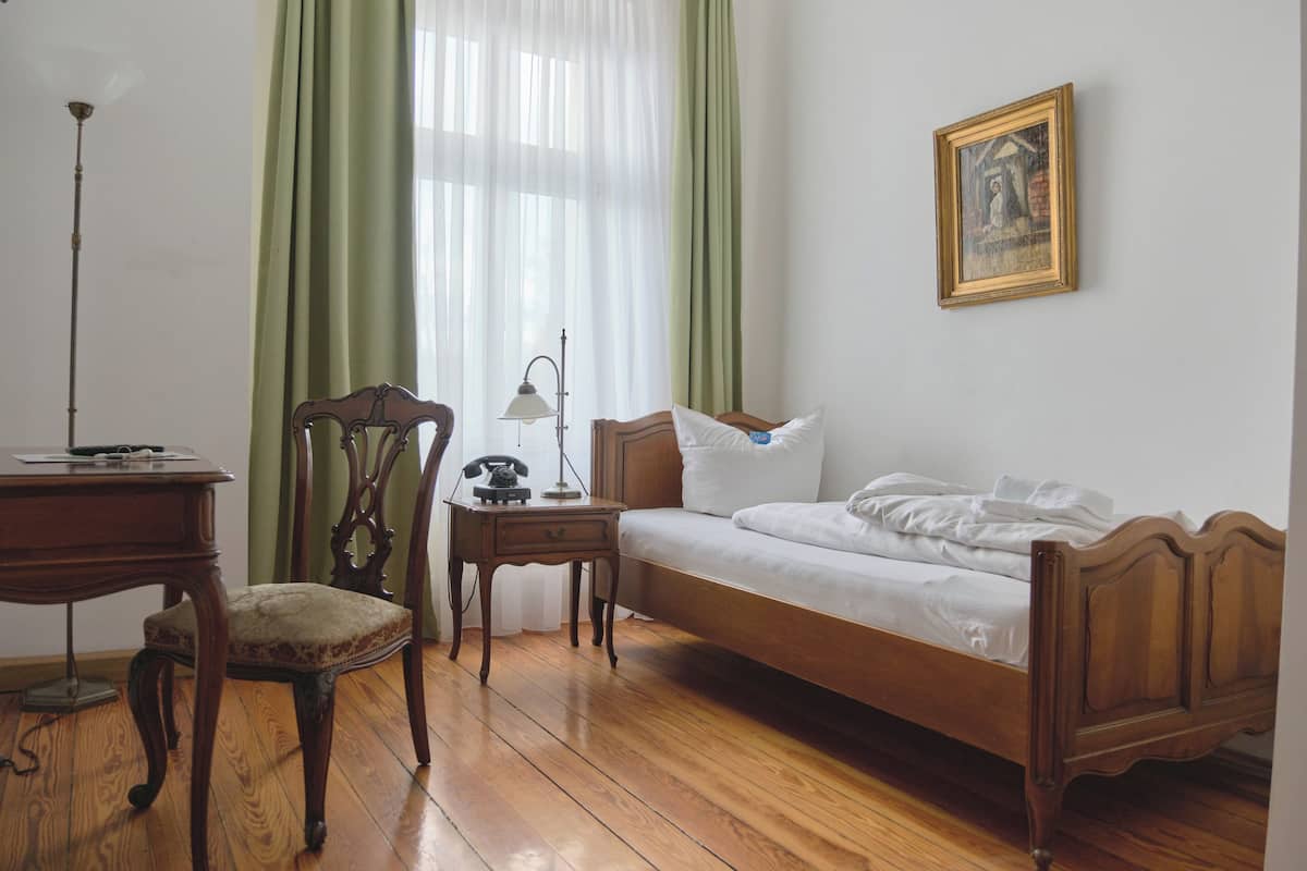 Quarto com cama de madeira, uma escrivaninha, cadeira, abajur e um quadro na parede.