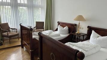 Quarto de hotel com duas camas de madeira, uma cadeira, uma mesa de cabeceira com abajur e uma janela grande com cortinas leves.