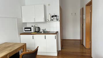 A compact kitchen with white cabinets, a microwave, a kettle, and a wooden dining table with a chair.