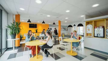 Un comedor moderno con mesas amarillas y blancas, lámparas colgantes negras y una planta grande en maceta.