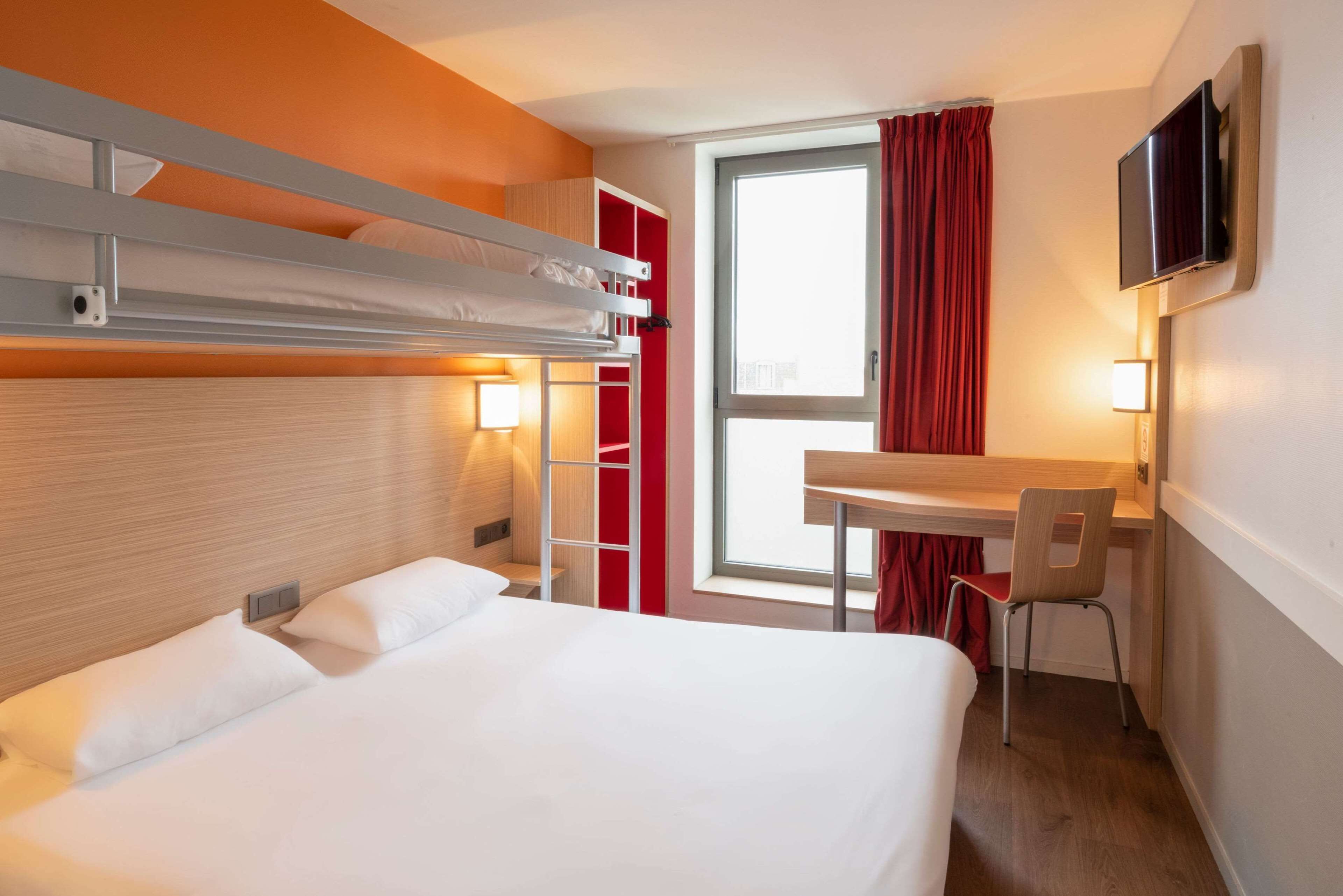 A hotel room with a bunk bed, a desk with a chair, a TV mounted on the wall, and a window with curtains.