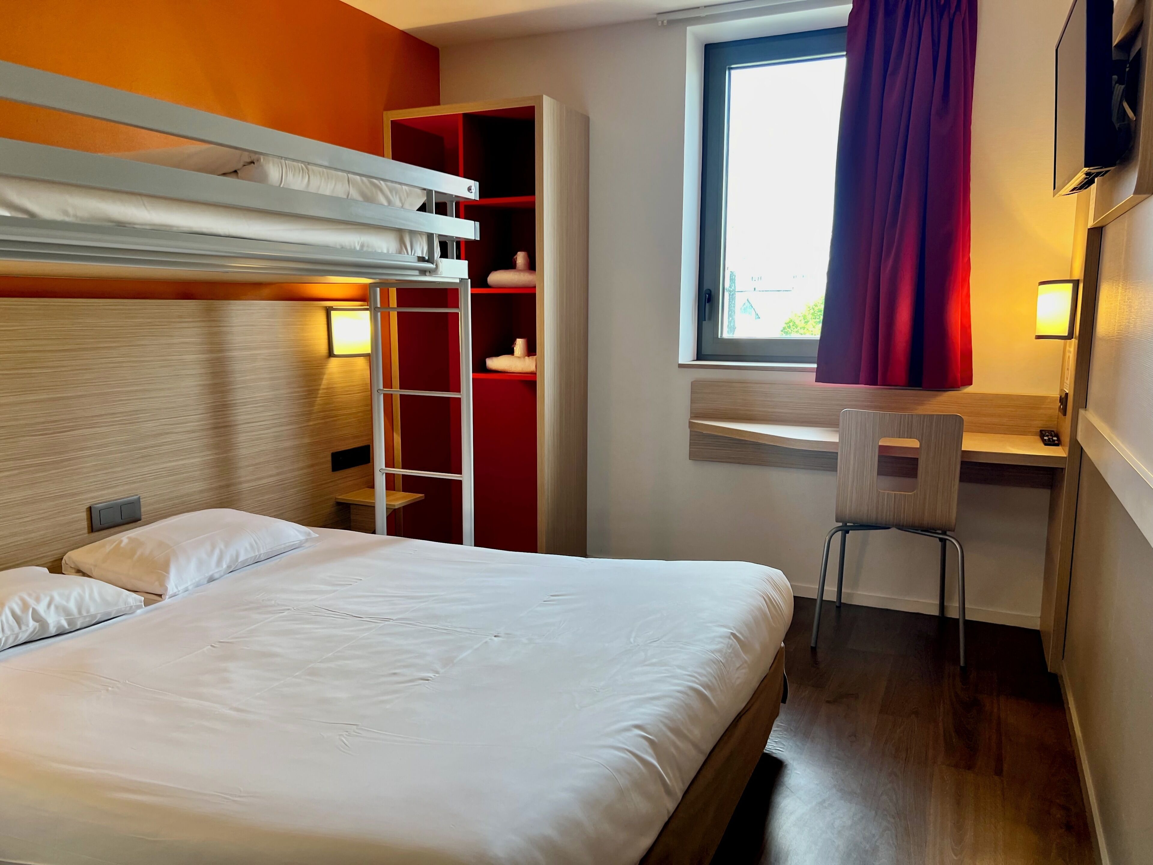 Una camera d'albergo con un letto a castello, una scrivania con sedia, un armadio e una finestra con tende.