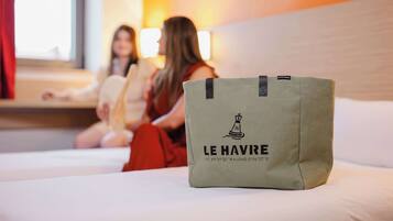 A hotel room with a bed, a green bag with the hotel name, and a window with curtains.