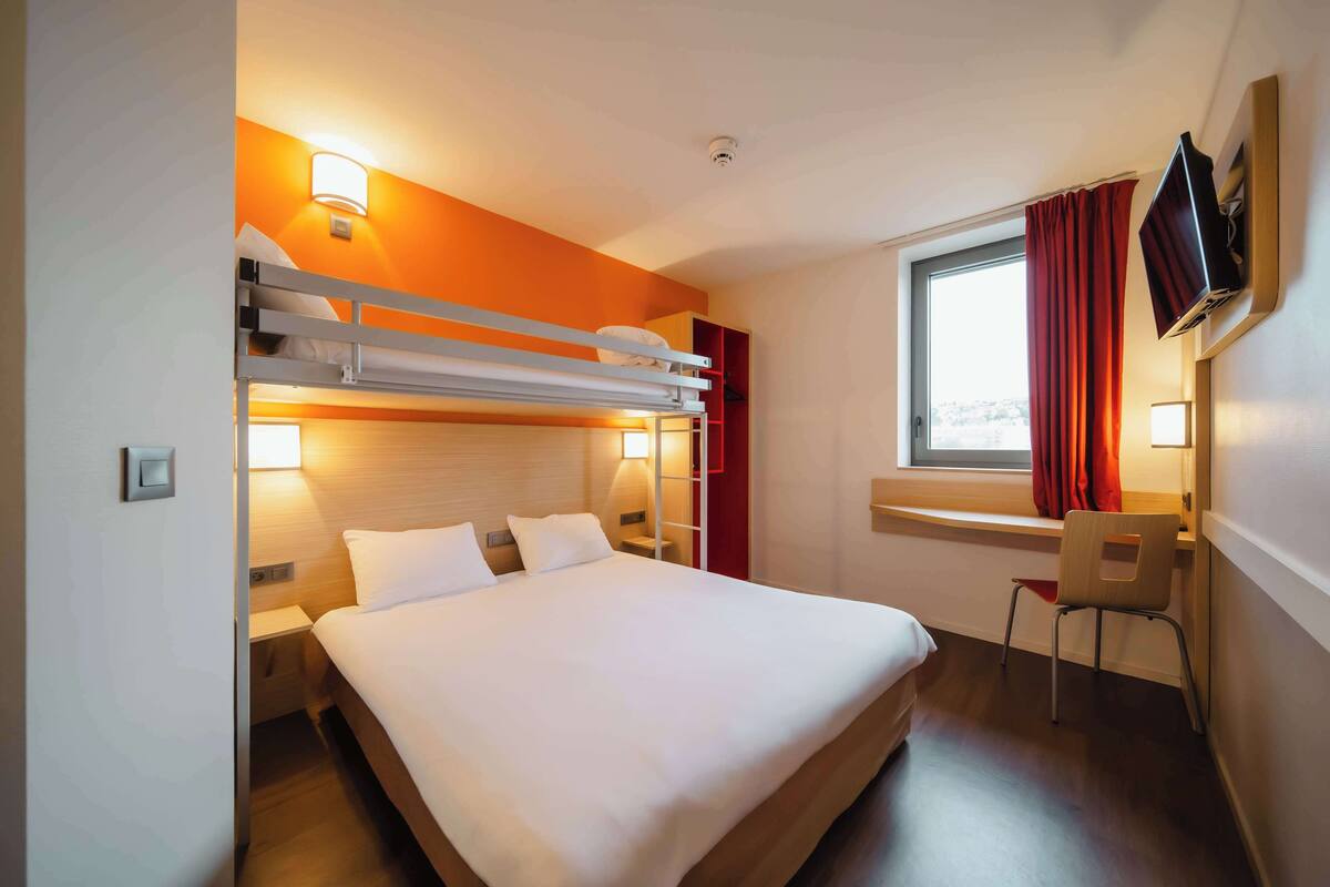 Habitación de hotel con literas, un escritorio con silla, televisor montado en la pared y ventana con cortinas.