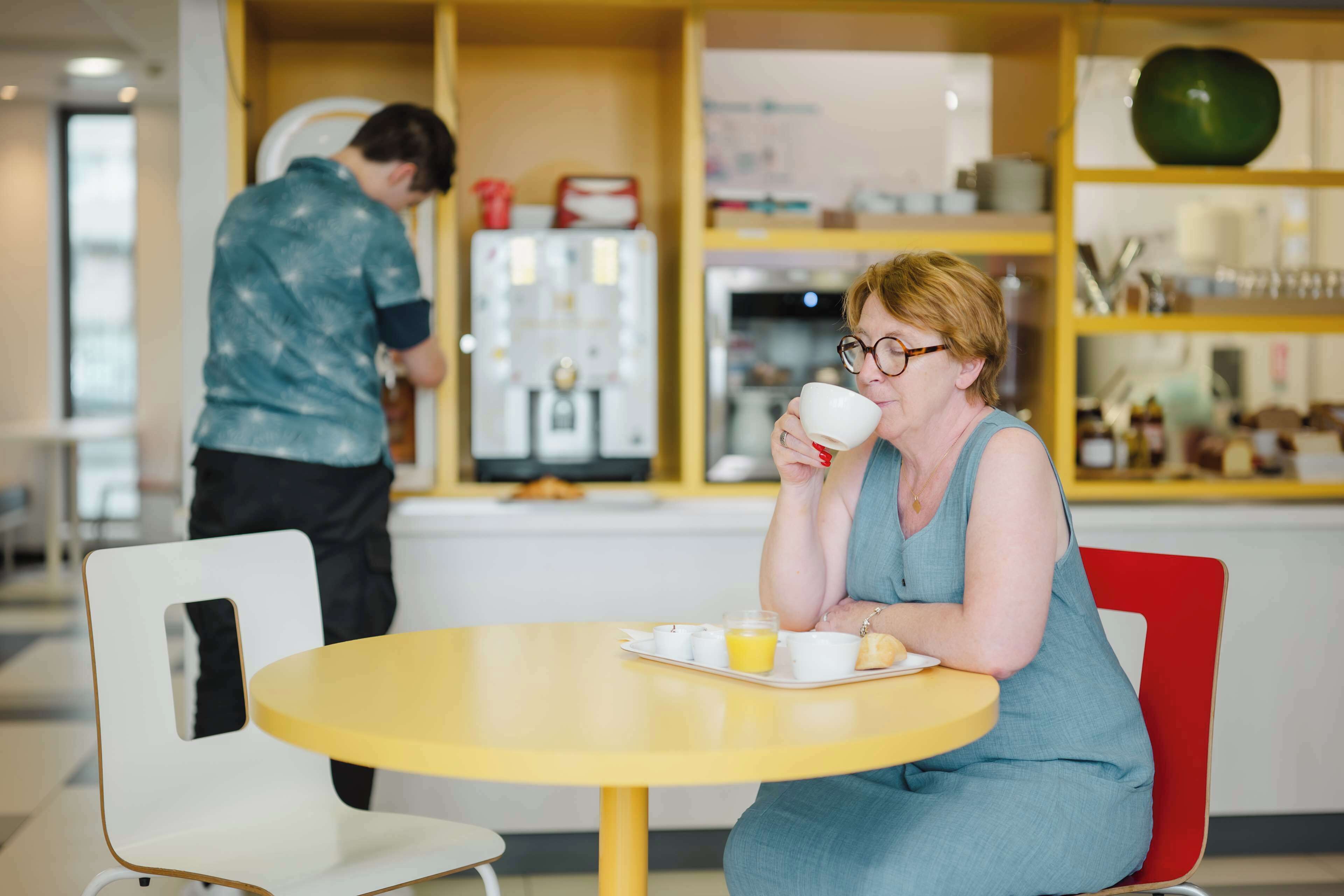 Una mujer sentada en una mesa amarilla en un café, sosteniendo una taza. Un hombre está de pie al fondo, y se ve una máquina de café.