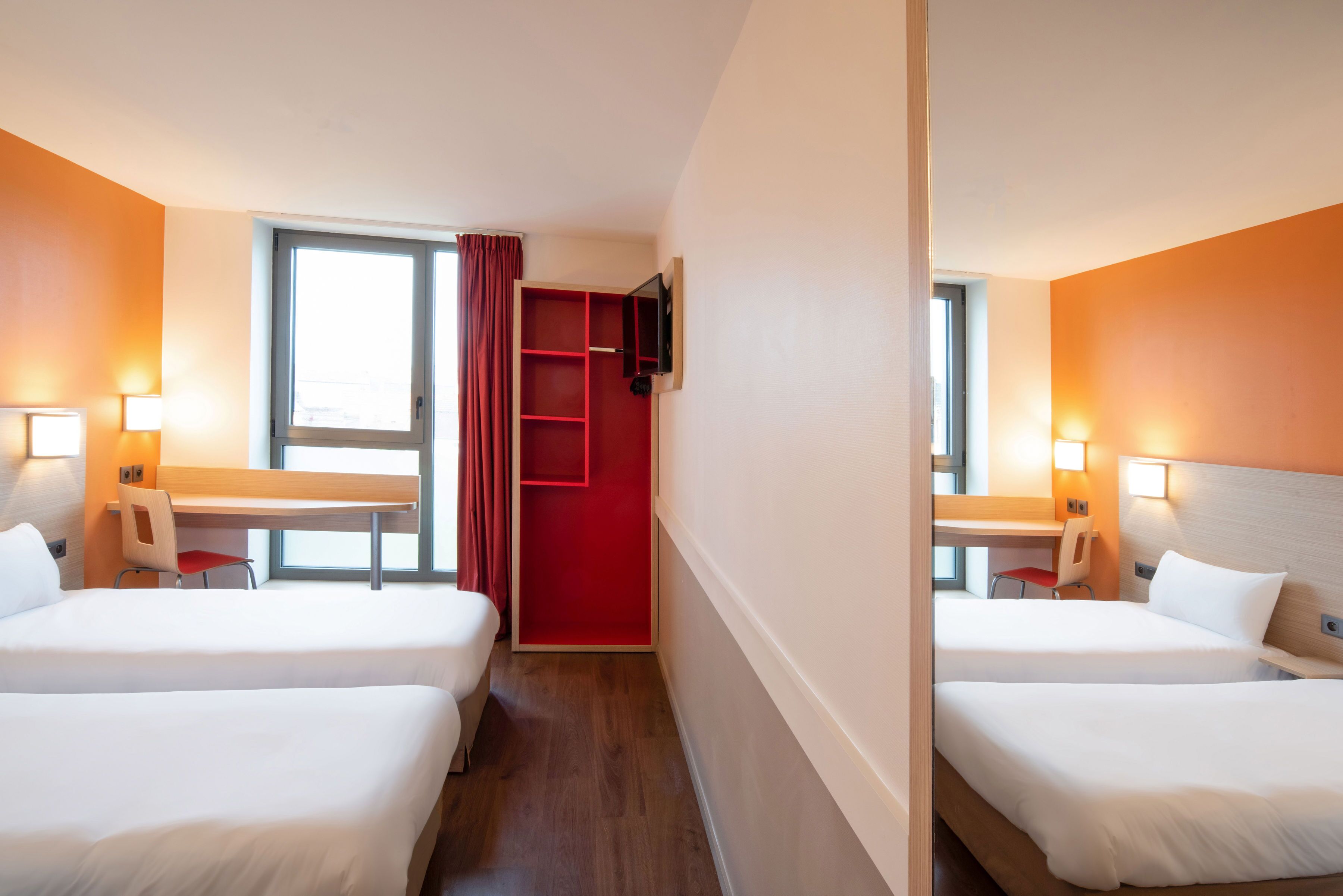 Une chambre d’hôtel avec deux lits, un bureau, une chaise, un téléviseur fixé au mur et une fenêtre avec des rideaux.