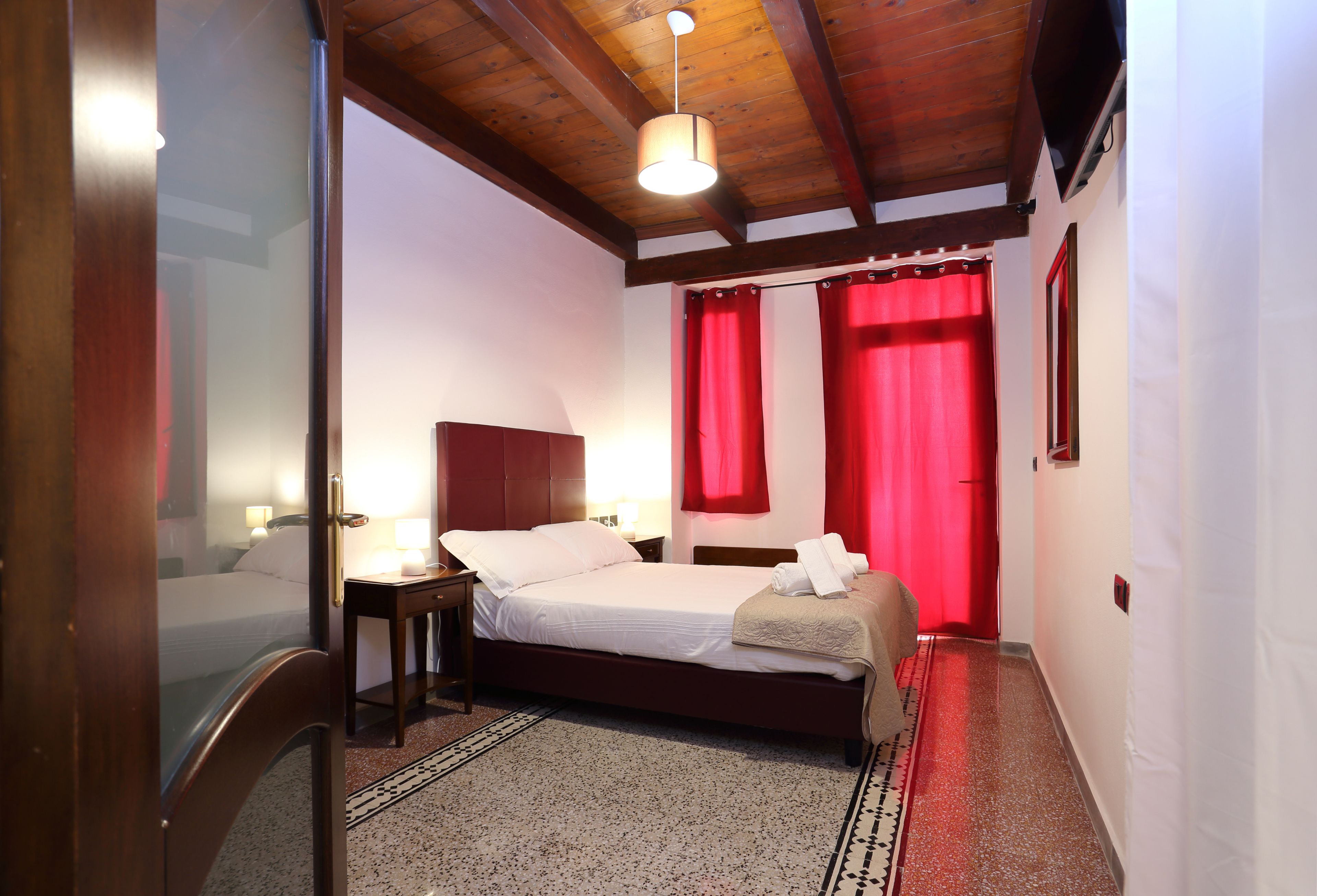 Ein Hotelzimmer mit Holzboden, einem Bett mit weißen Leinen, einem Nachttisch mit Lampe und roten Vorhängen.