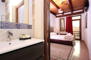 Camera d'albergo con bagno dotato di lavandino bianco, specchio e schienale piastrellato. La stanza presenta un letto con tenda rossa, un comodino con lampada e vista da una finestra.