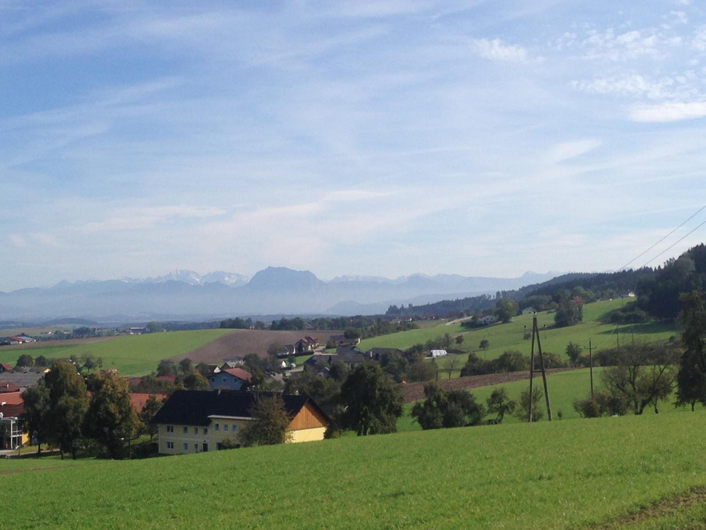 Un paysage rural avec des maisons sur une colline, un ciel dégagé et des montagnes à l’horizon.