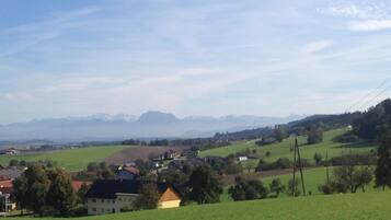 Un paysage rural avec des maisons sur une colline, un ciel dégagé et des montagnes à l’horizon.