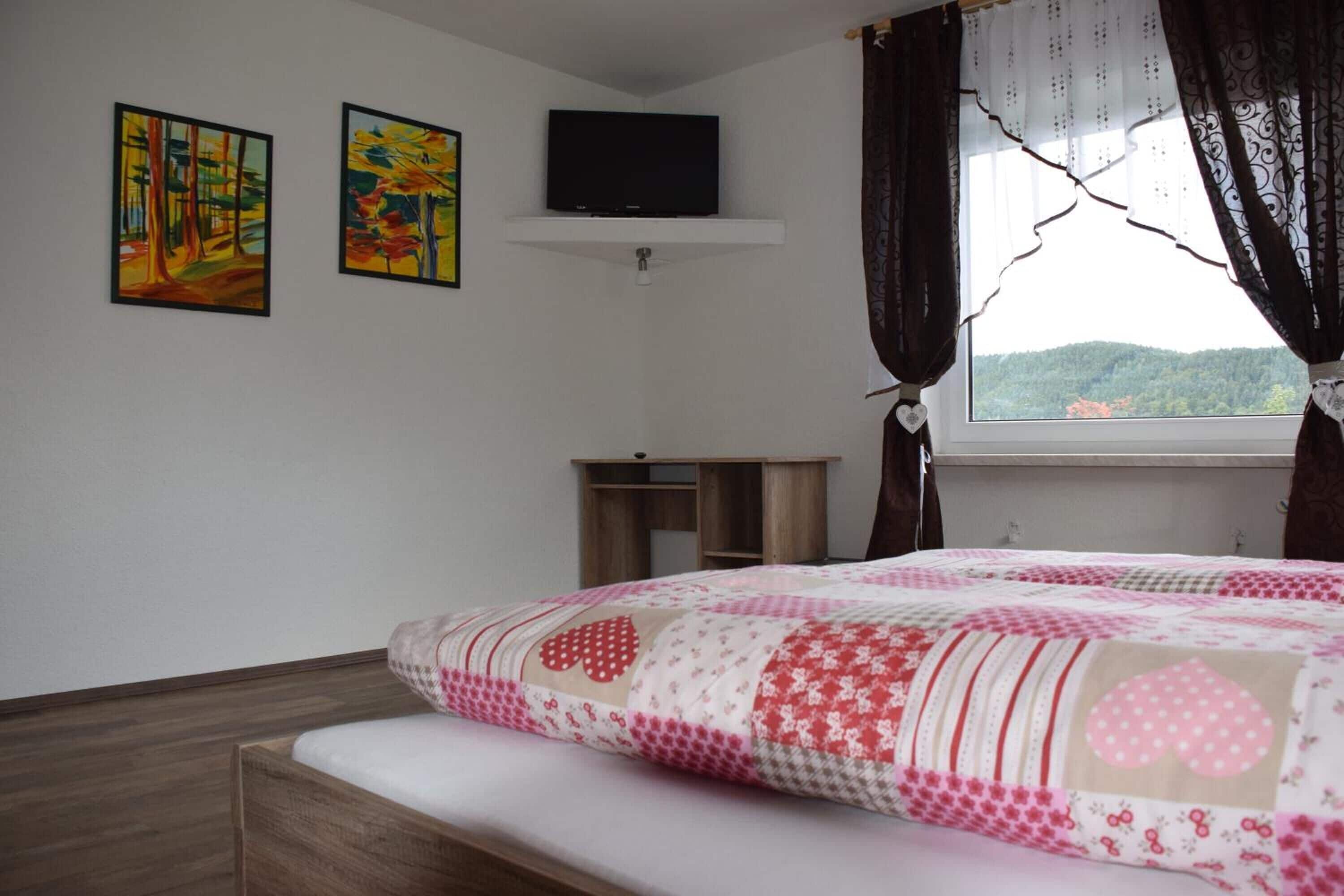 Un dormitorio con cama, televisor en estante, dos cuadros en la pared y ventana con cortinas.