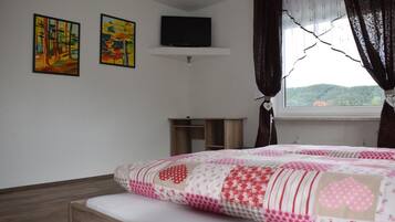 Un dormitorio con cama, televisor en estante, dos cuadros en la pared y ventana con cortinas.
