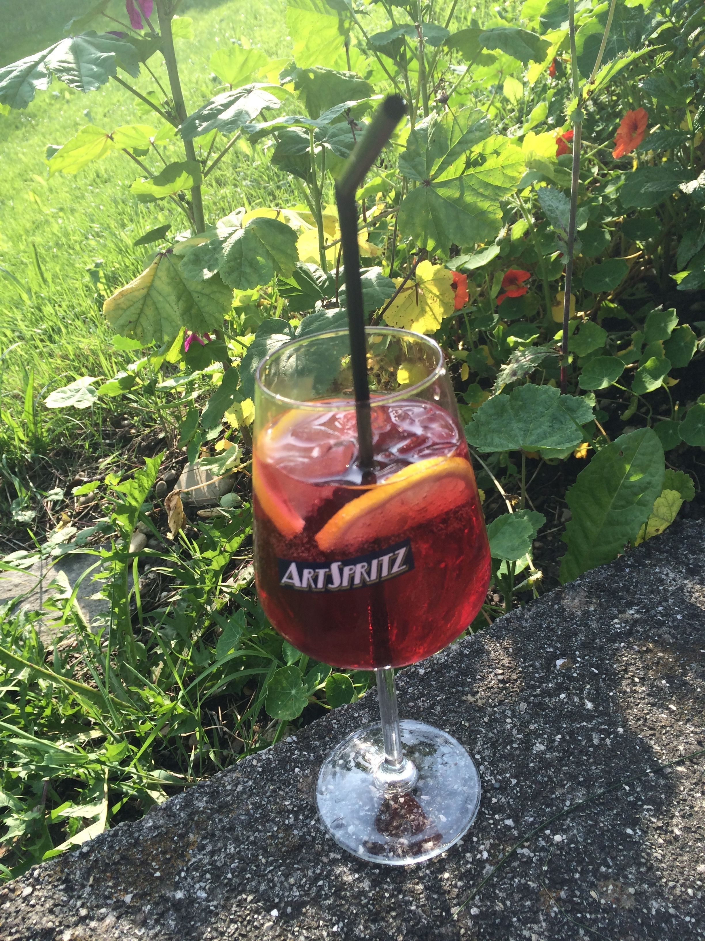 A glass of red beverage with a straw, labeled 'ARTSPRITZ', on a stone surface outdoors.