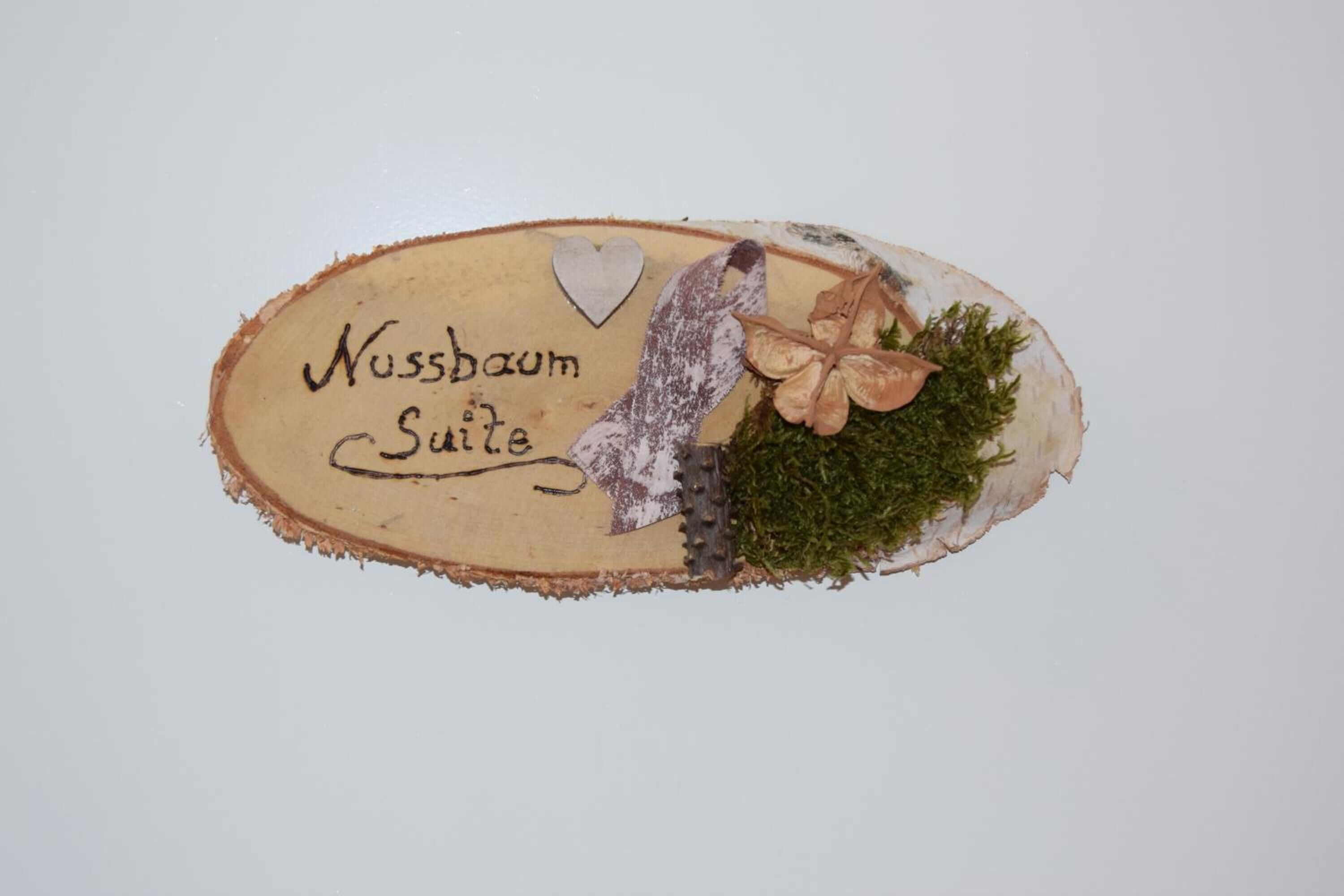 Eine hölzerne Plakette mit dem Schriftzug 'Nussbaum Suite' und dekorativen Elementen, darunter ein Herz und Blätter.