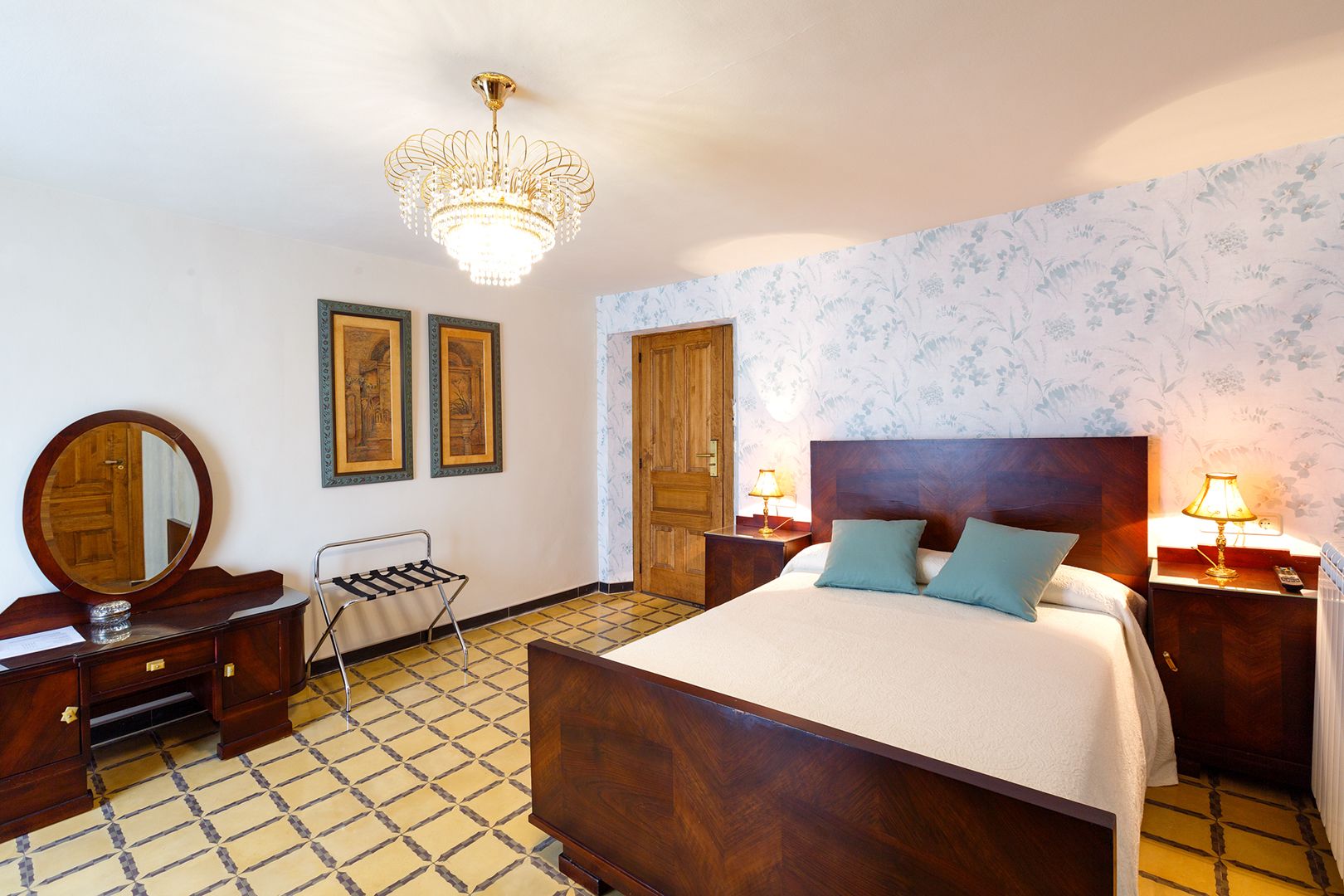 Habitación de hotel con una cama grande, cabecera de madera, dos mesitas de noche con lámparas, cómoda de madera, espejo, una silla y una araña.