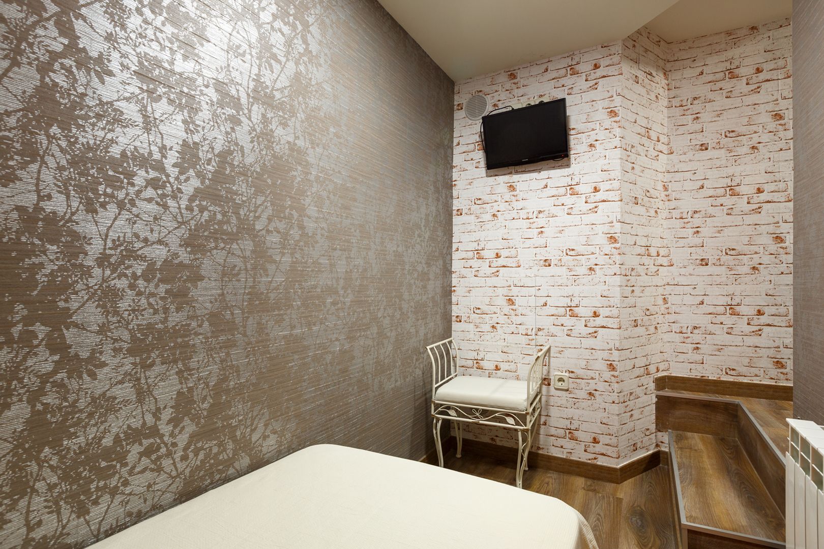 Une petite pièce comprenant un lit, une chaise, un téléviseur fixé au mur, qui est décoré d’un motif en briques, et un plancher en bois.