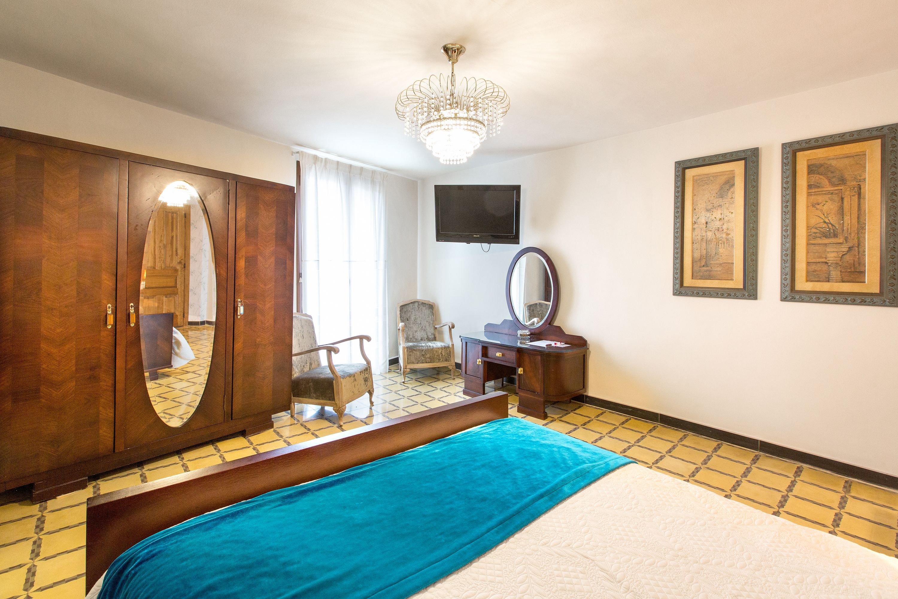 A hotel room with a large bed, a wooden wardrobe, a desk with a mirror, a television on the wall, and two framed pictures.