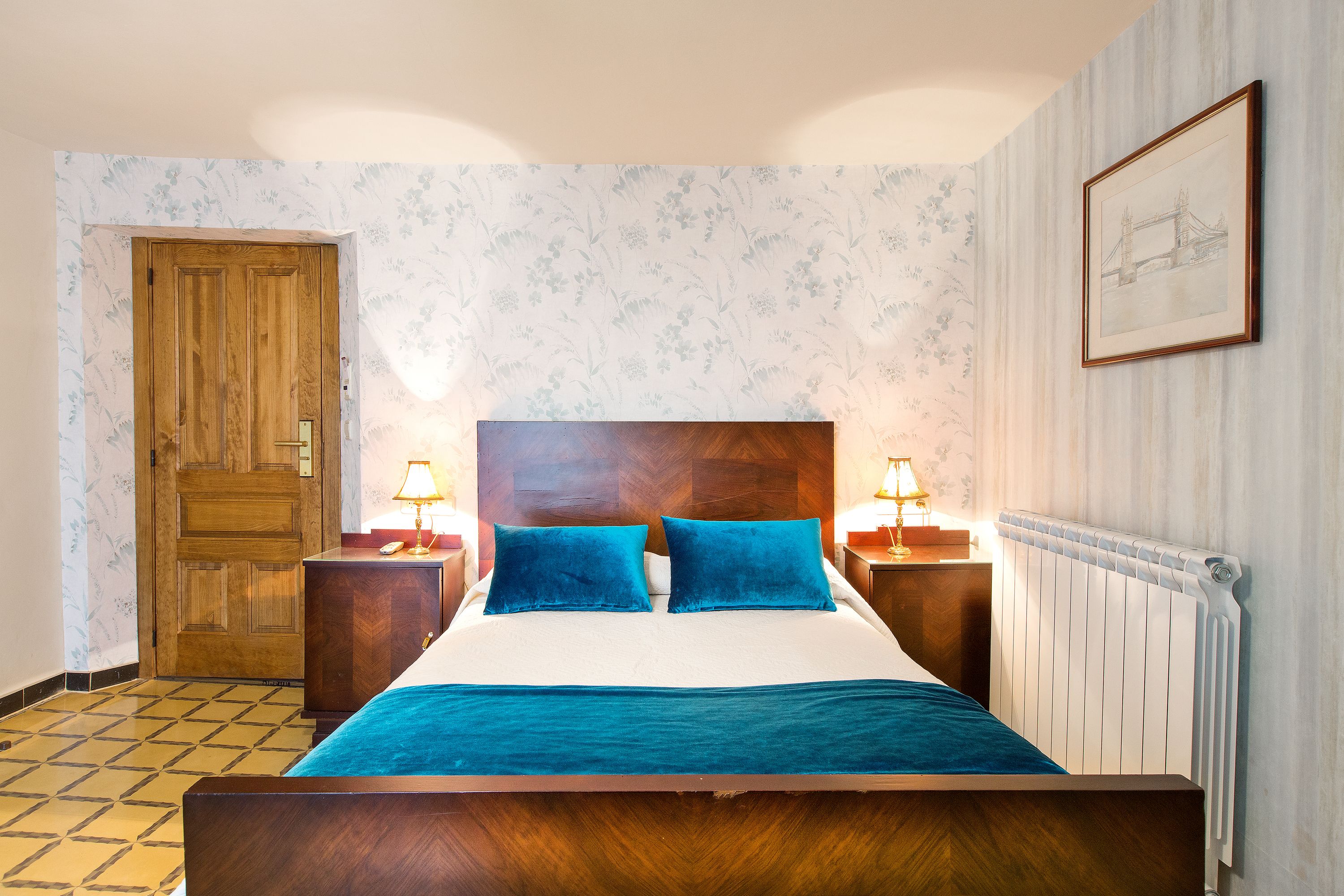 Un dormitorio con cama de madera, dos almohadas azules, edredón blanco, cabecero de madera, dos mesitas de noche con lámparas, un armario de madera, una puerta y un radiador.