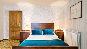 Un dormitorio con cama de madera, dos almohadas azules, edredón blanco, cabecero de madera, dos mesitas de noche con lámparas, un armario de madera, una puerta y un radiador.