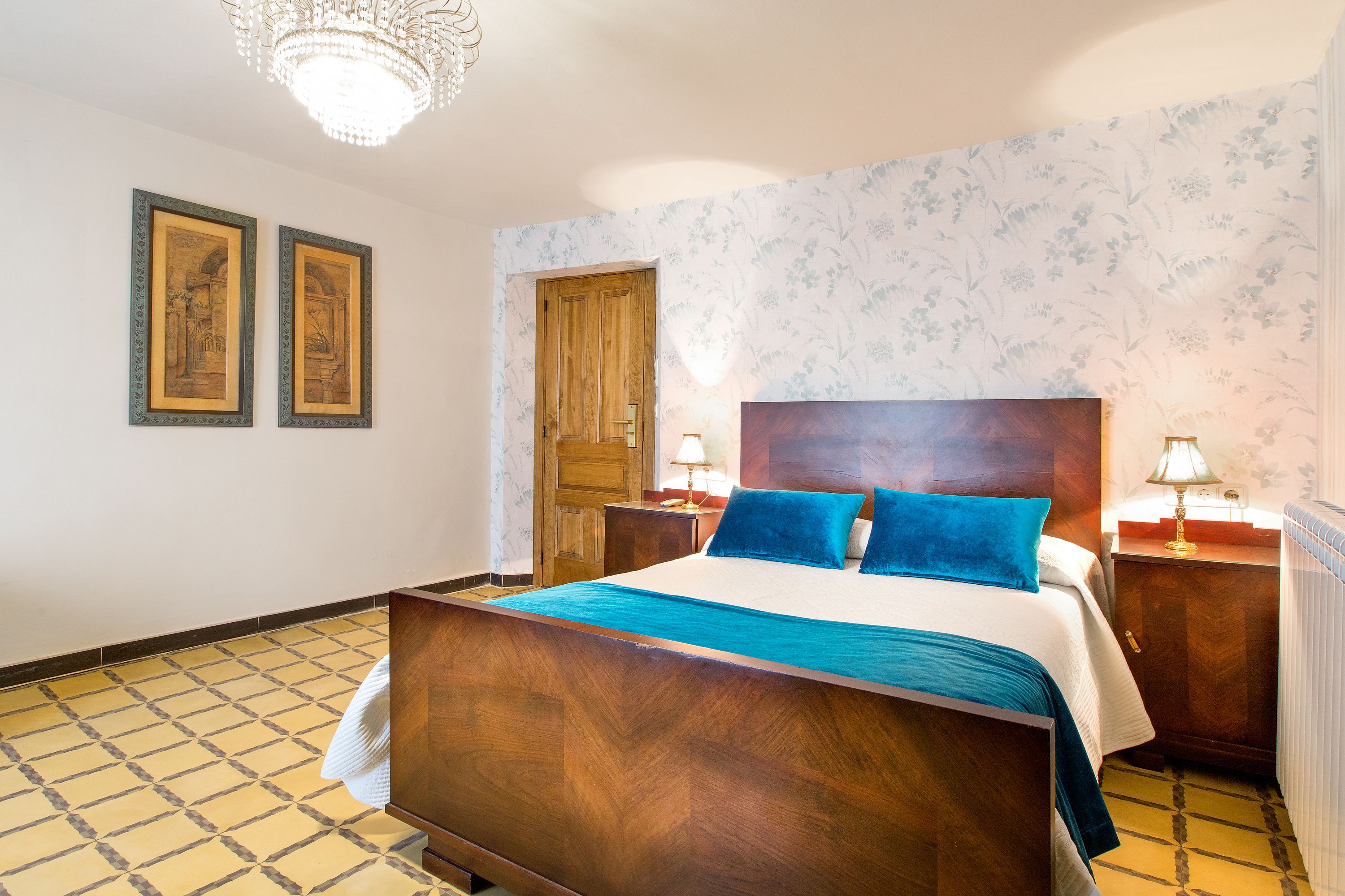 A hotel room with a wooden bed, two framed pictures on the wall, a wooden door, and a patterned wallpaper.