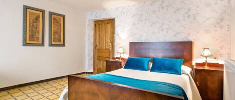 Habitación de hotel con cama de madera, dos cuadros enmarcados en la pared, una puerta de madera y papel tapiz estampado.