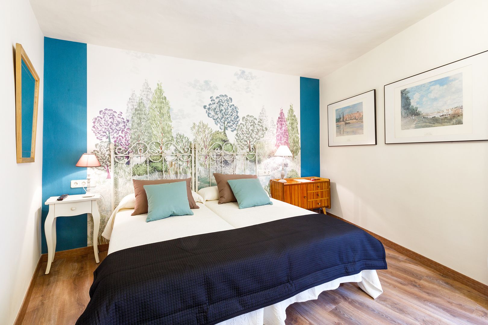 Un dormitorio con una cama, dos mesitas de noche, una mesita auxiliar, un mural en la pared y dos cuadros enmarcados.