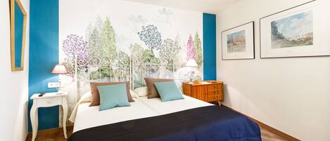 Un dormitorio con una cama, dos mesitas de noche, una mesita auxiliar, un mural en la pared y dos cuadros enmarcados.