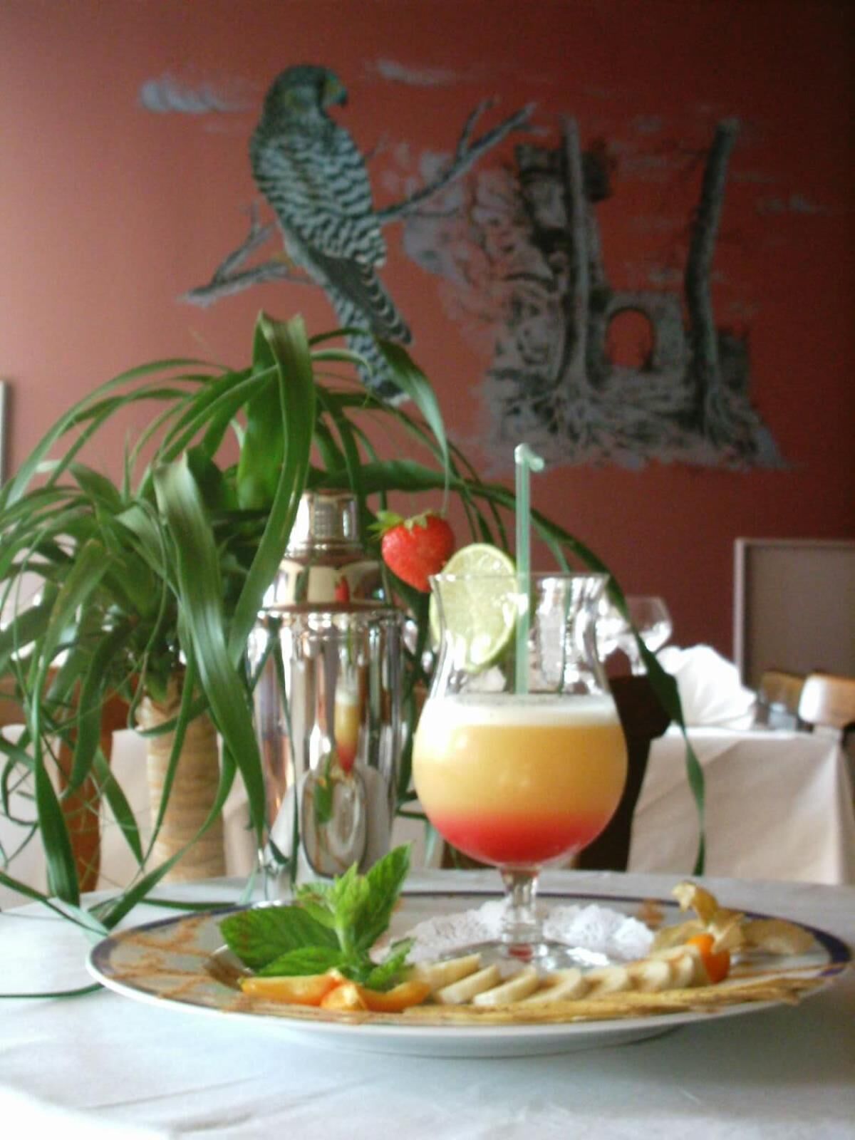 A table with a drink, a plate of food, a cocktail shaker, and a painting in the background.