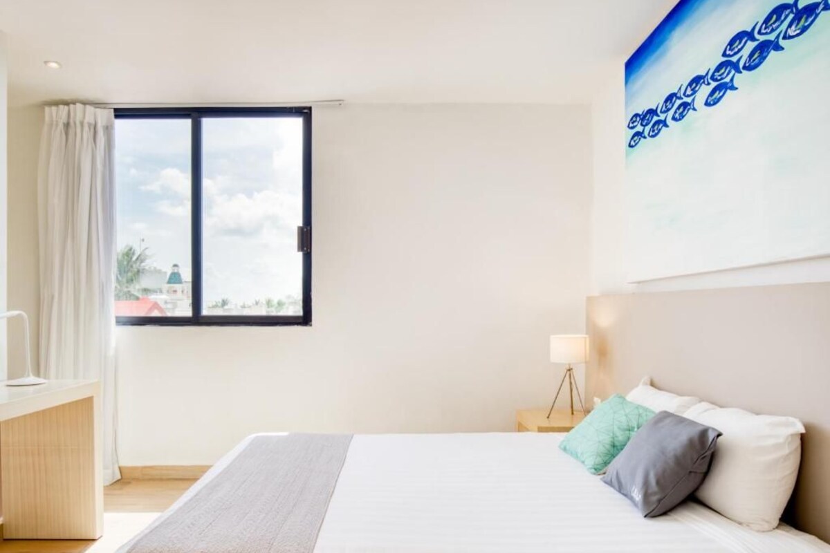 A hotel room with a bed, a desk, a lamp, and a window with a view of a building and trees.