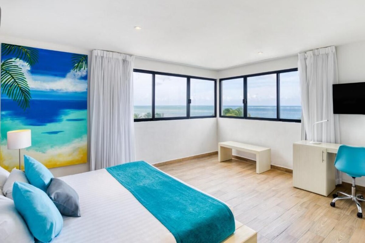 Habitación de hotel con una cama grande, un escritorio, una silla, un banco y un cuadro de una playa.