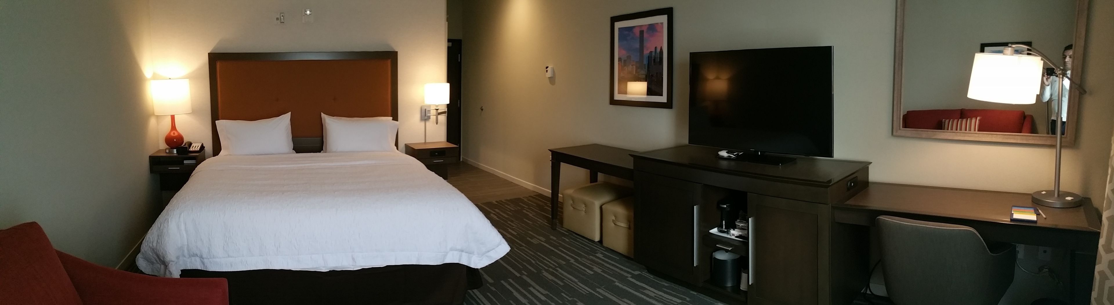 Una habitación de hotel con una cama grande, un televisor, un escritorio con lámpara y un pequeño cubo de basura.