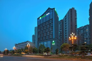A multi-story building with a Holiday Inn Express sign, surrounded by other high-rise buildings and street lamps. - Holiday Inn Express Leshan City Square by IHG (Leshan)