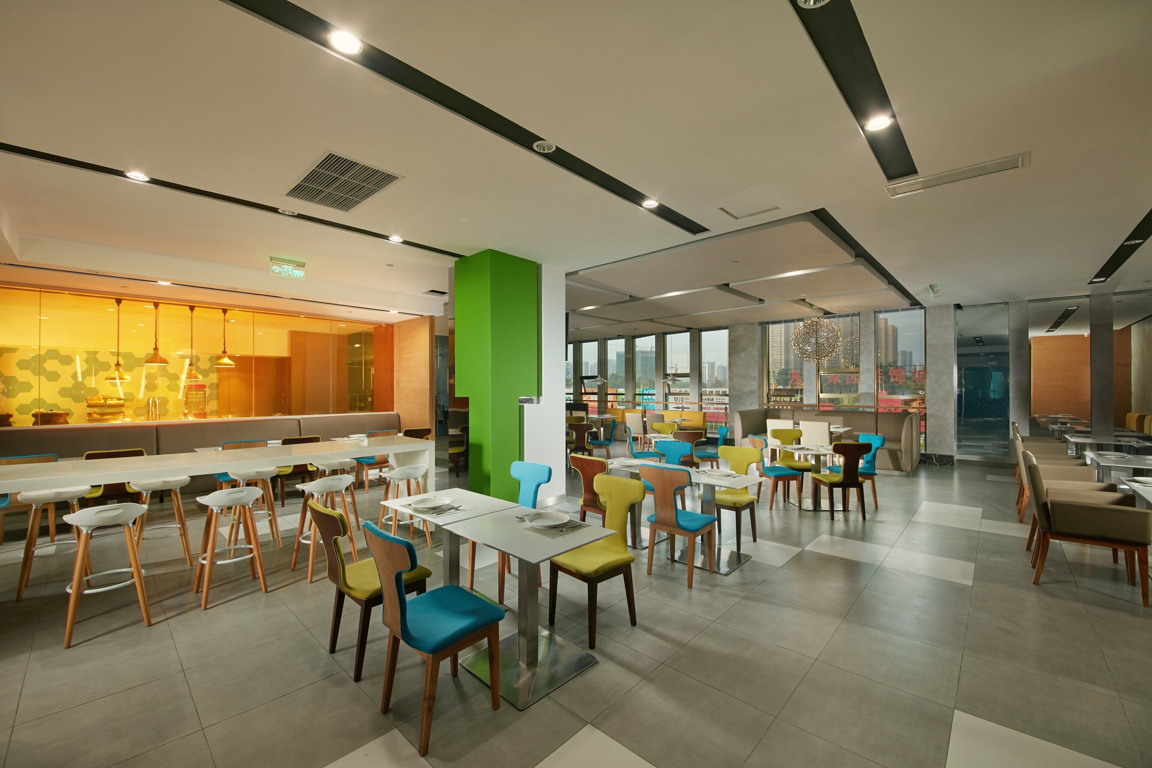 Una cafetería moderna con varias mesas y sillas, una pared de acento verde y grandes ventanales con vista a la ciudad.