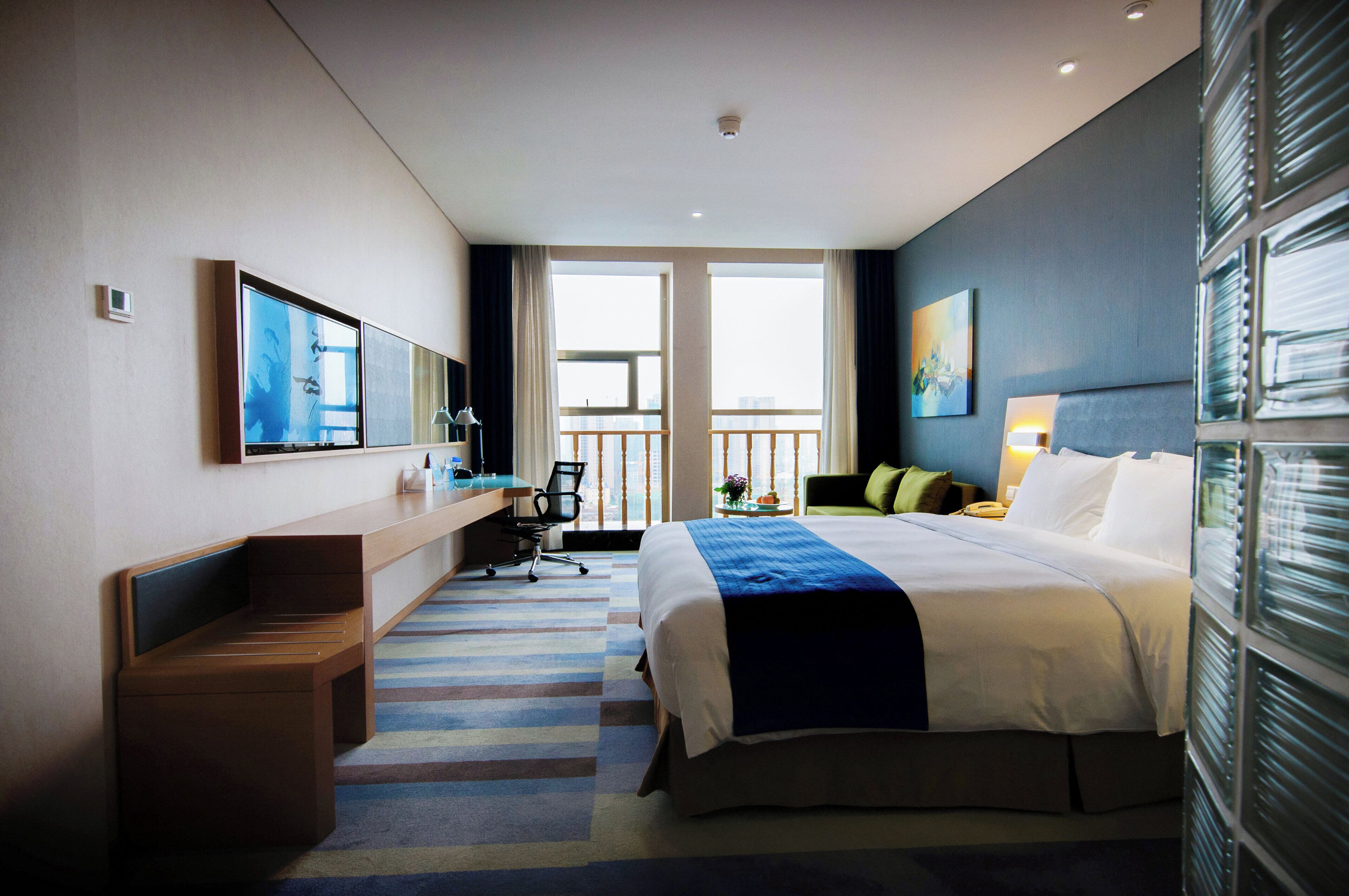 Une chambre d’hôtel équipée d’un lit, d’un bureau, d’une chaise et offrant une vue sur la ville grâce à de grandes fenêtres.